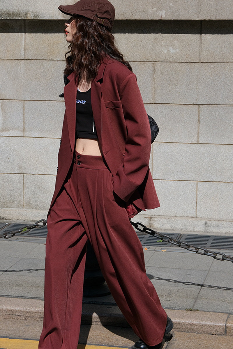 Model wearing a burgundy wide-leg suit set with a relaxed-fit blazer and high-waisted trousers from Belle Vestige. Styled with a cropped black top and a casual cap, showcasing a bold and confident look perfect for modern, empowered women.